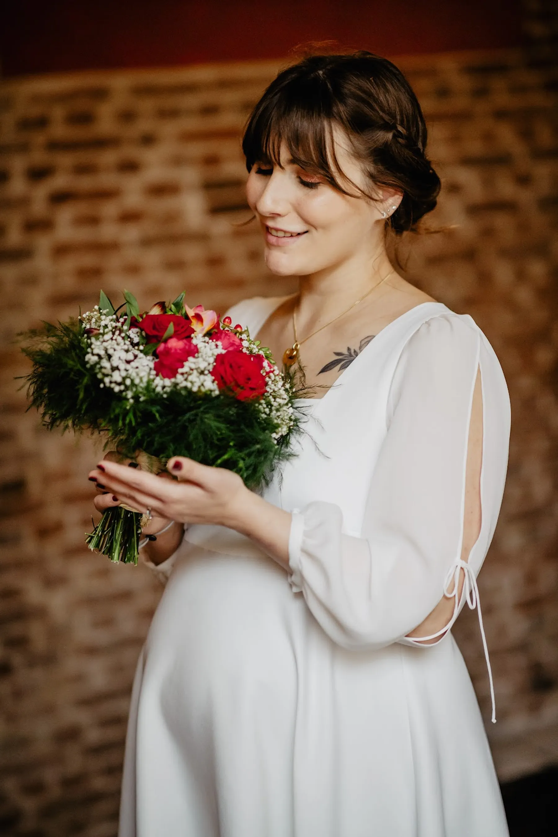 Adelina en robe de mariée sur mesure mi-longue à manches fendues nouées, bouquet de roses rouges et gypsophile à la main