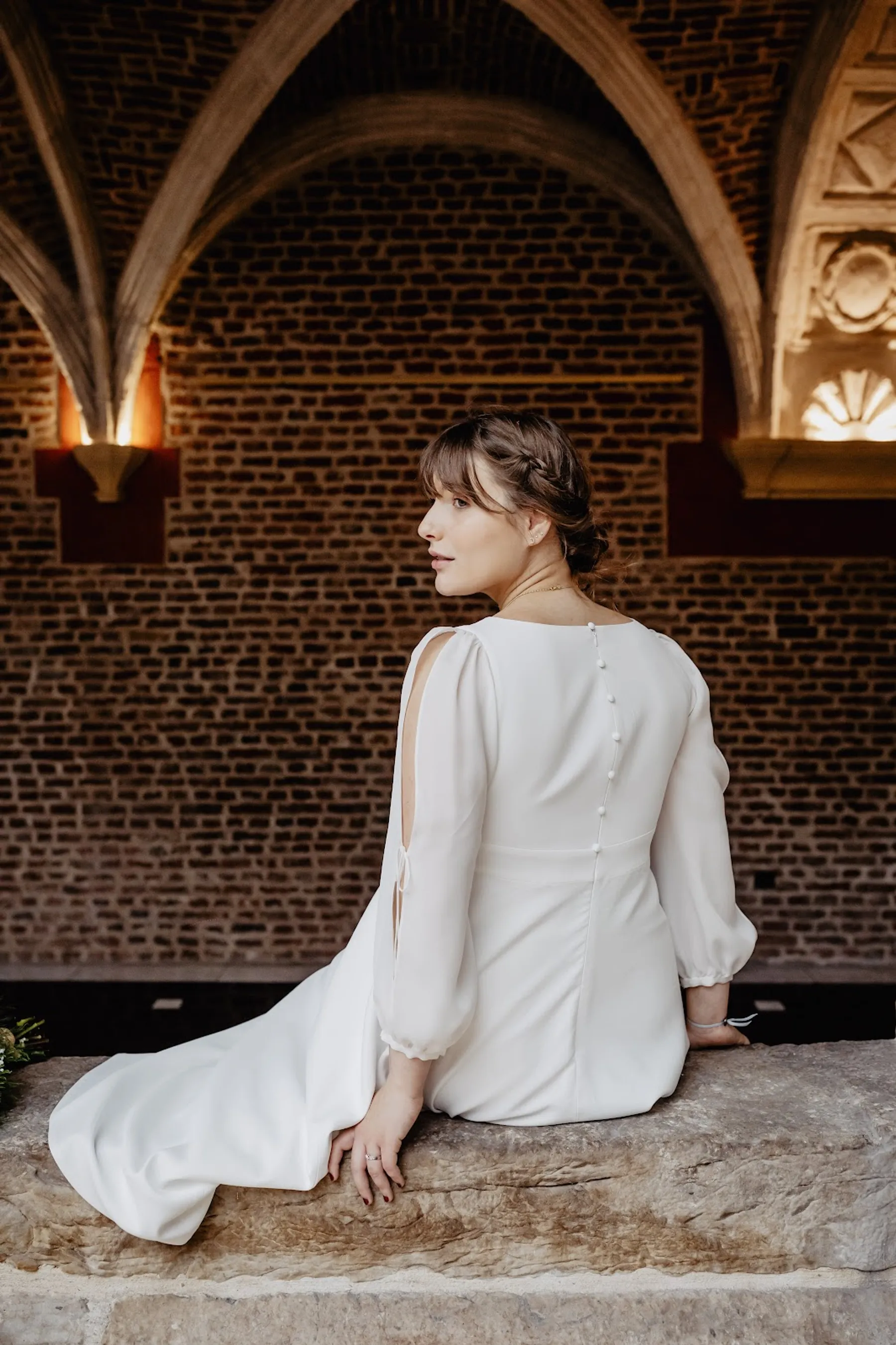 Dos de la robe de mariée mi-longue à boutons couverts et manche fendue, mariée assise sur une pierre sous des voûtes en briques