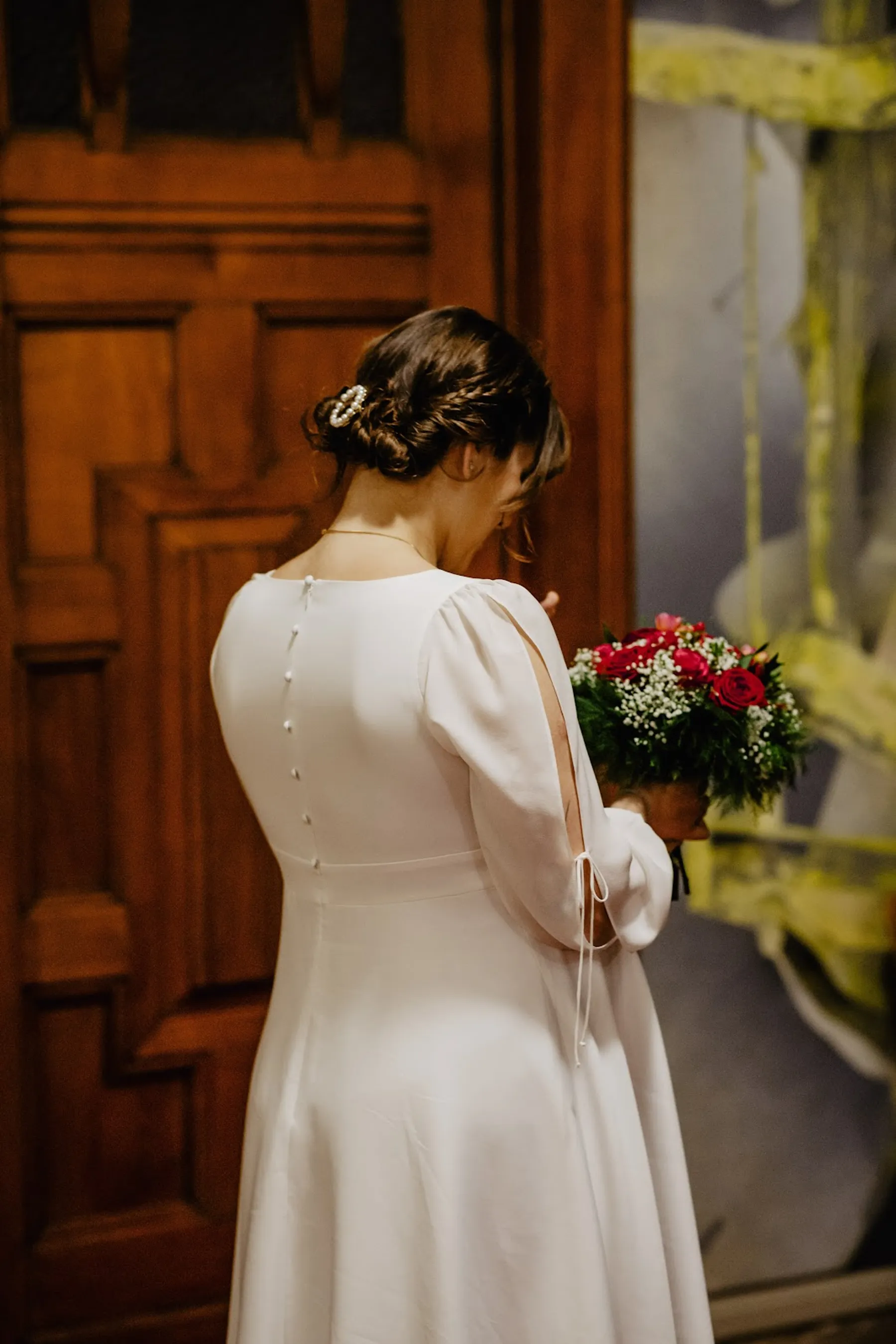 Adelina de dos devant une porte en bois, robe de mariée à boutons couverts et manche fendue, bouquet de roses rouges