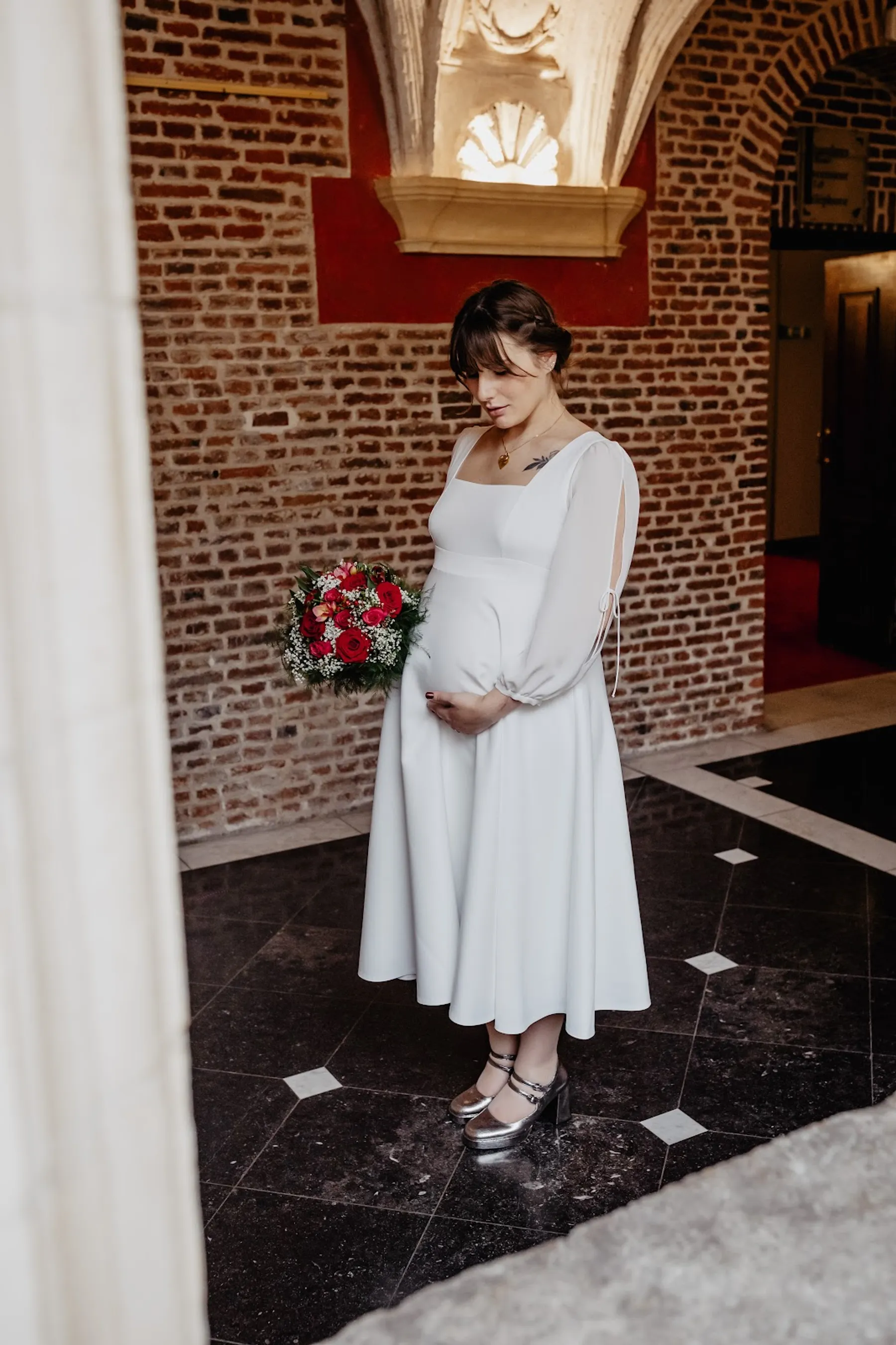 Adelina debout sous une voûte en briques, robe de mariée mi-longue à col carré et manches fendues, bouquet de roses rouges