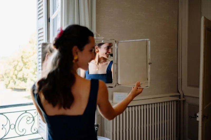 Agathe se prépare devant un petit miroir, de dos, en robe d'invitée sur mesure bleu nuit à larges bretelles et col carré