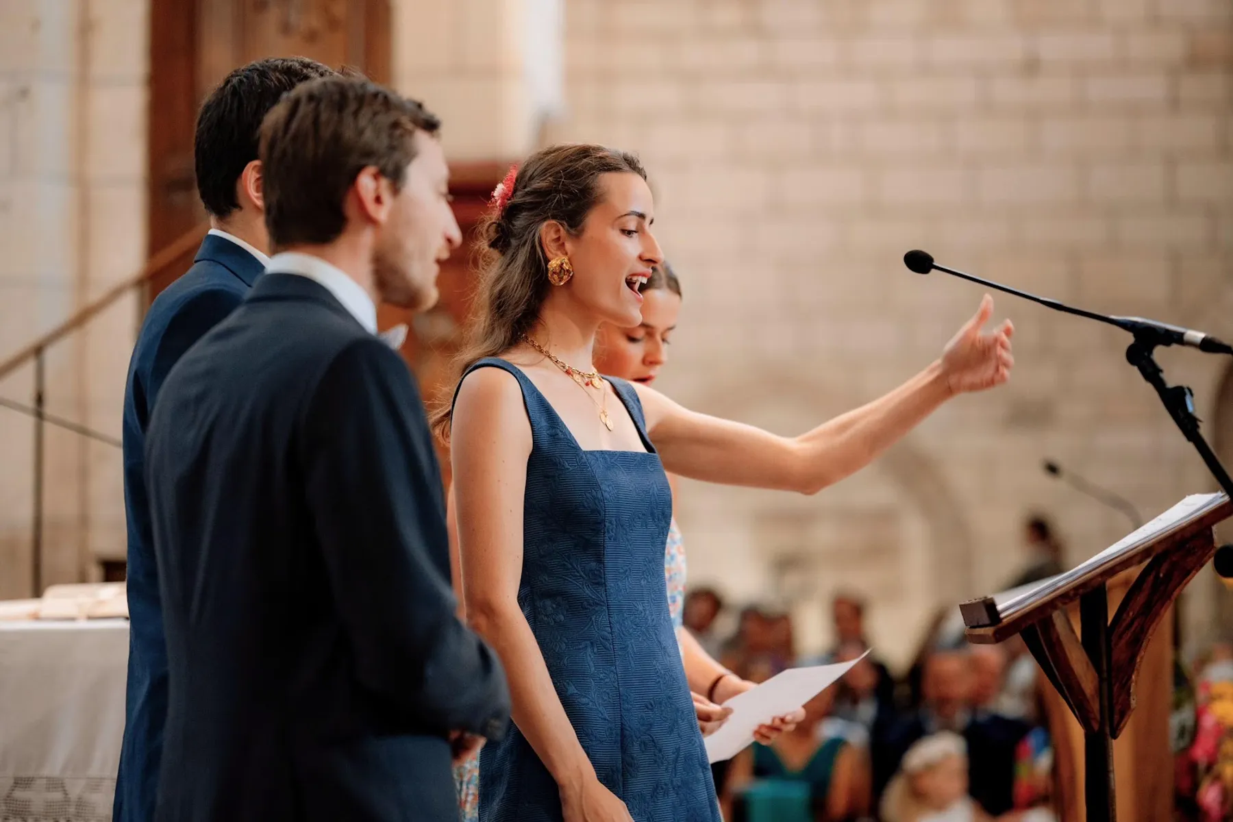 Agathe lit un texte au micro pendant la cérémonie, robe d'invitée bleue à col carré, une fleur rouge dans les cheveux