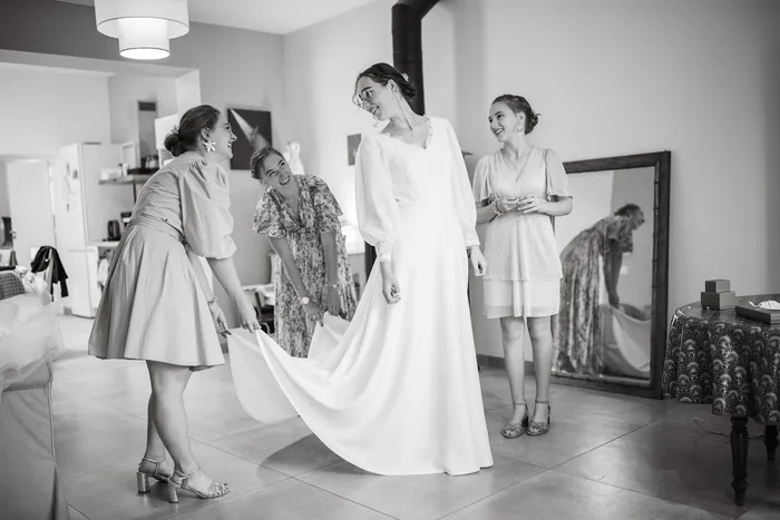 Alice en robe de mariée sur mesure à col V et longues manches, entourée de proches qui soulèvent sa traîne, en noir et blanc