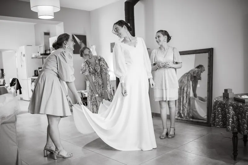 Alice en robe de mariée sur mesure à col V et longues manches, entourée de proches qui soulèvent sa traîne, en noir et blanc