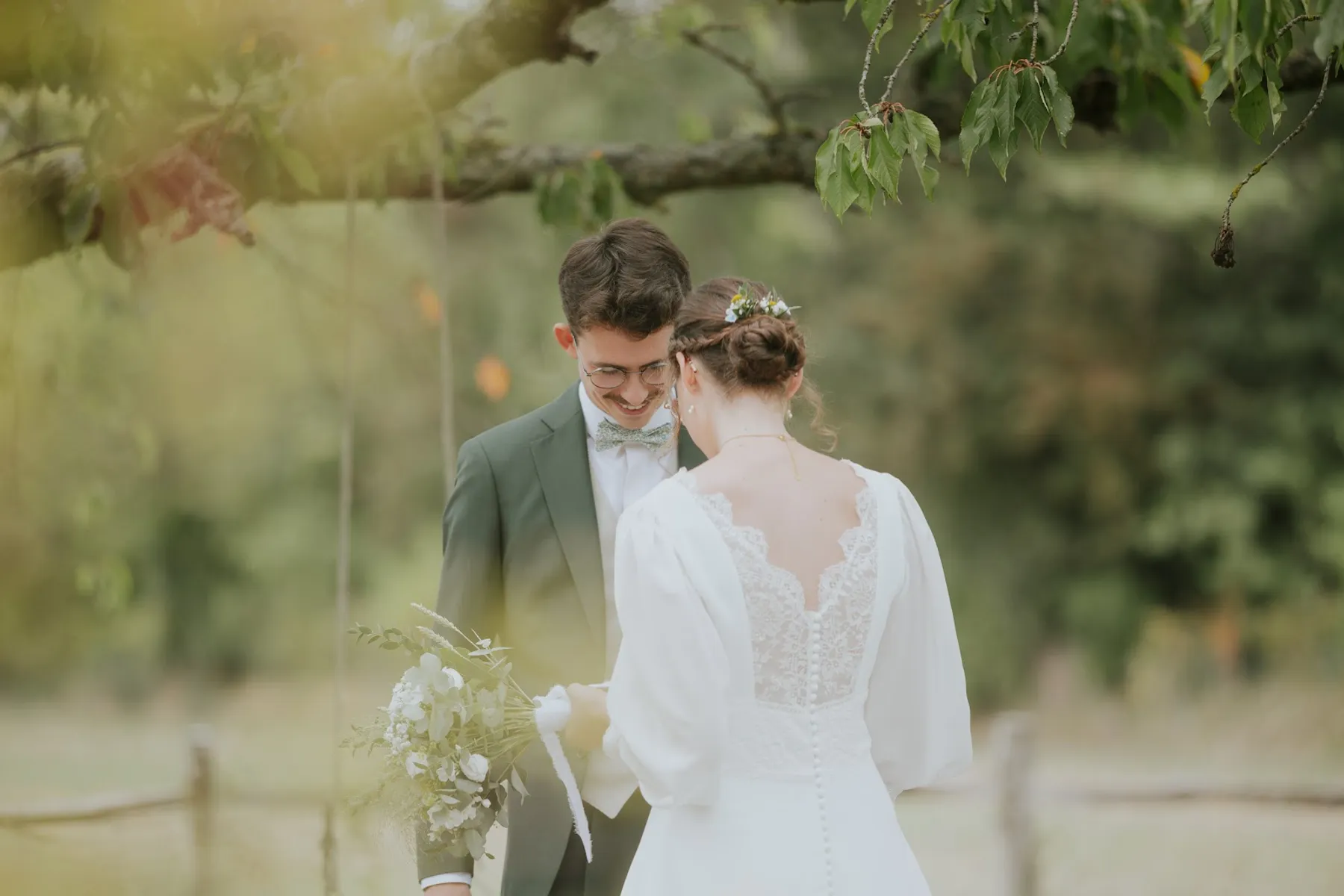 Dos de la robe de mariée d'Alice, décolleté en V en dentelle et rangée de boutons, sous un arbre avec son marié en costume vert