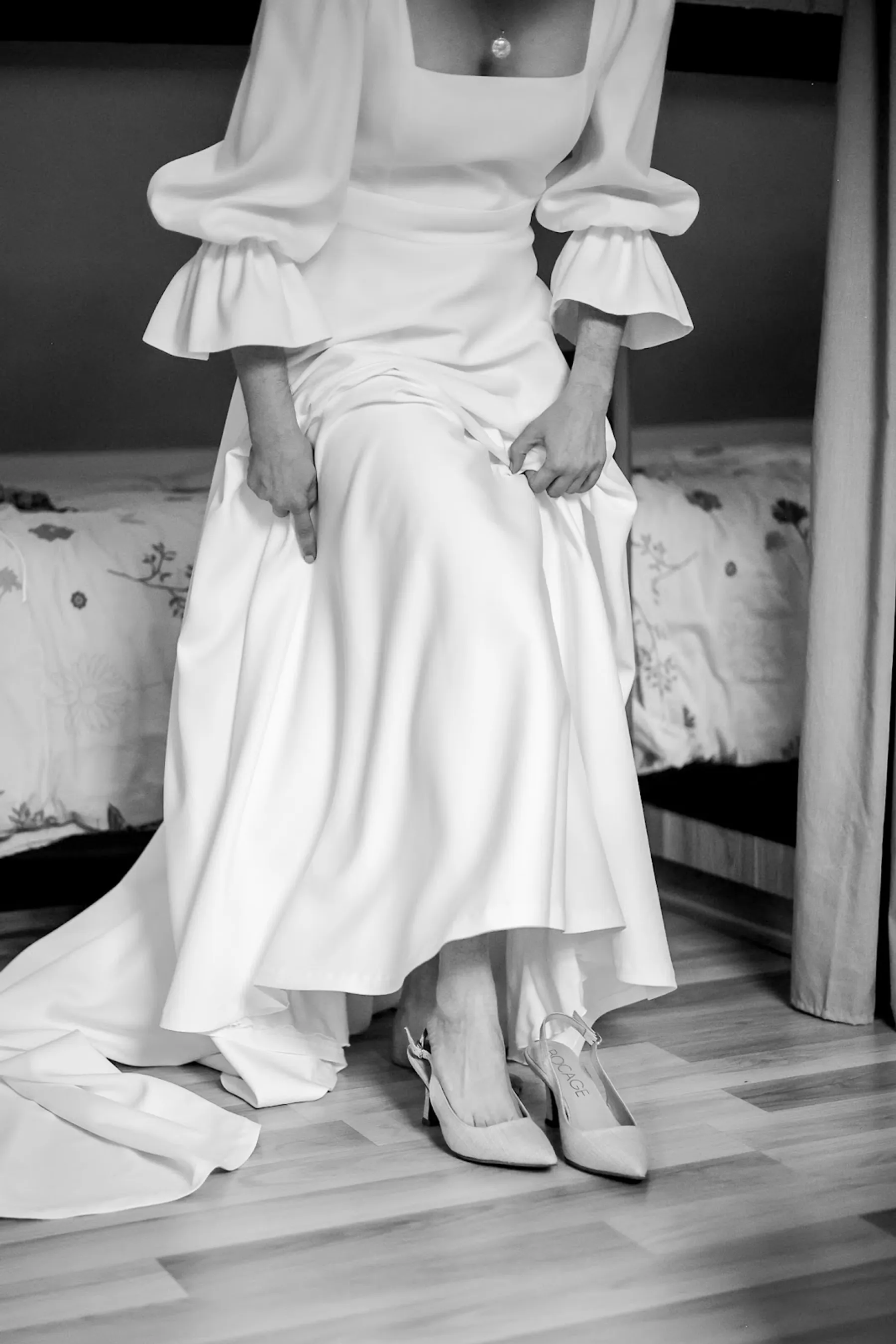 Mariée assise dans sa robe à col carré et manches à volants, jupe fluide et traîne étalée, escarpins aux pieds