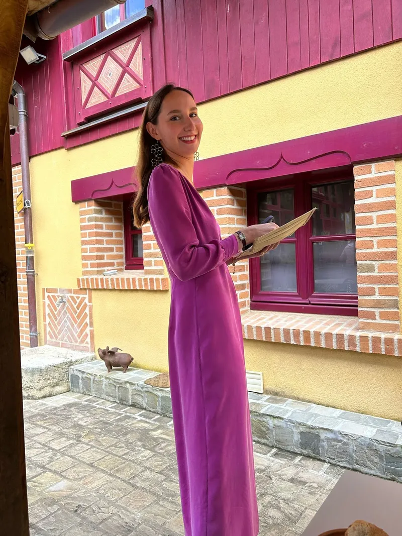 Auriane souriante en longue robe d'invitée violette à manches longues, devant une façade jaune et briques