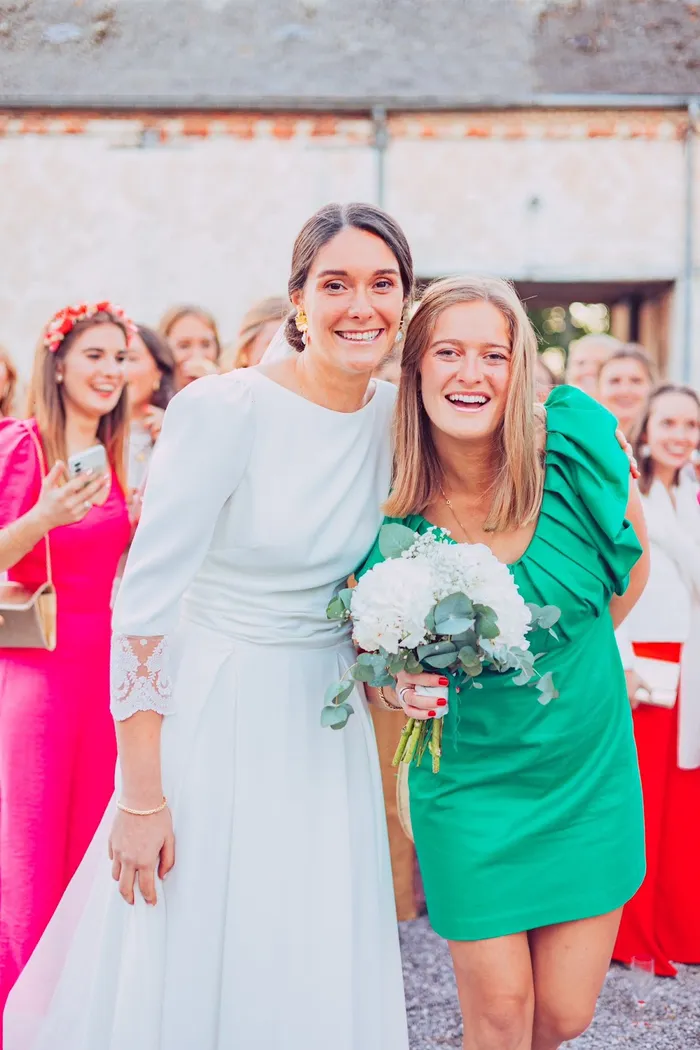Blanche en robe d'invitée verte à épaule volantée, serrée contre la mariée au milieu des invités
