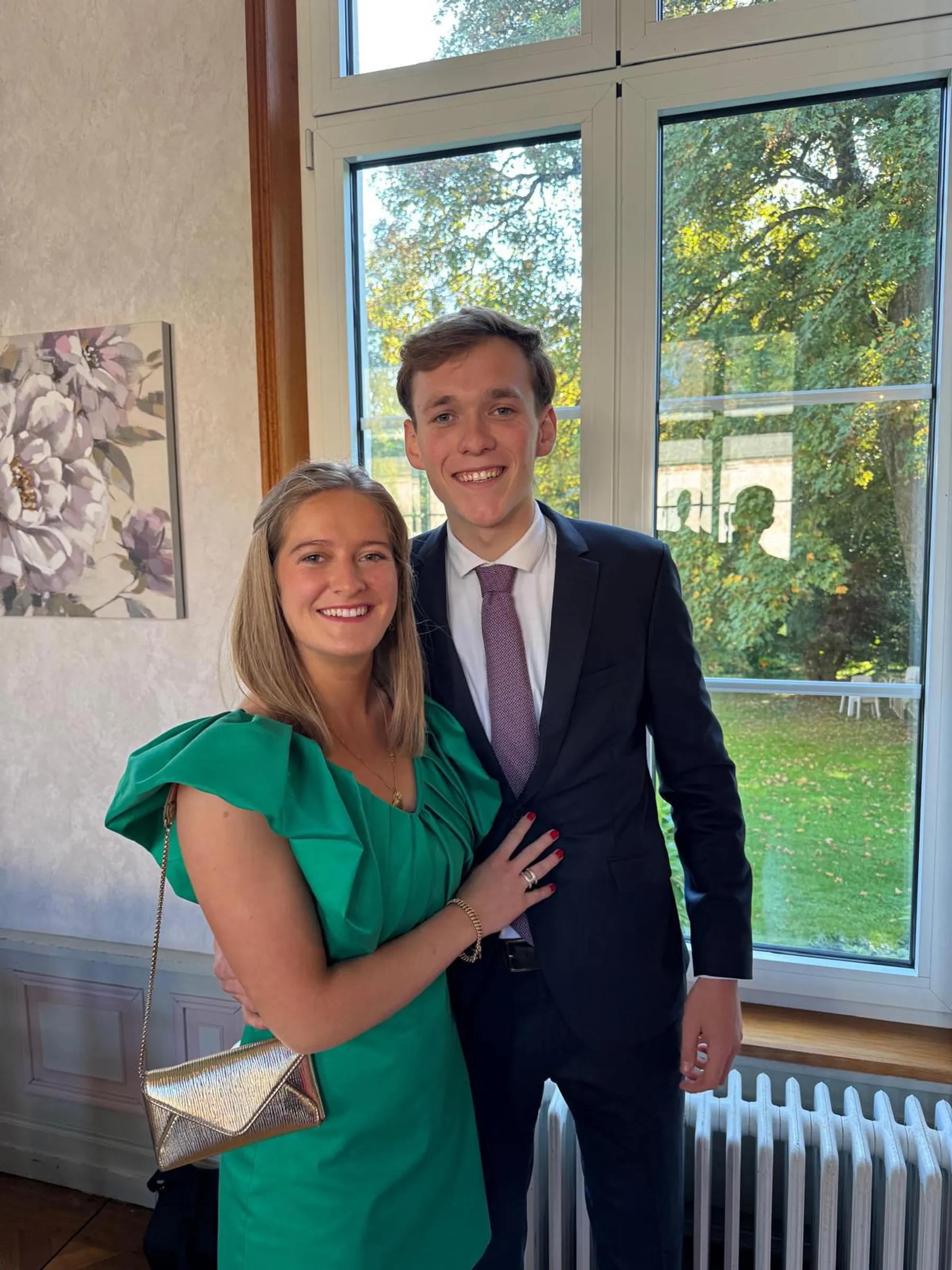 Blanche en robe verte à manche volantée et pochette dorée, dans une salle lumineuse, au côté d'un invité en costume