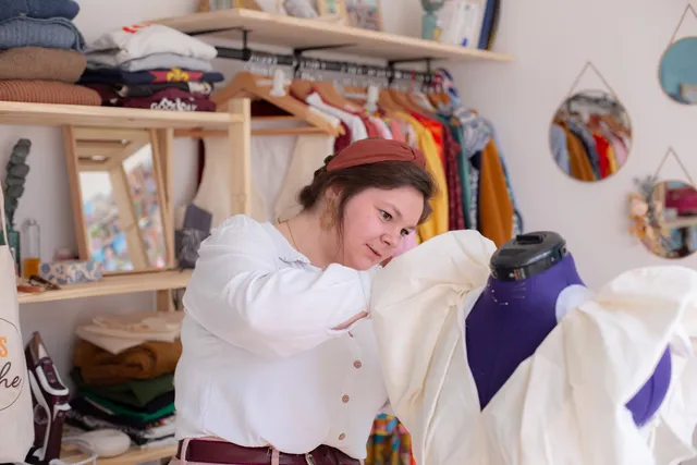 Hermine dans son atelier, entourée de vêtements et de tissus.