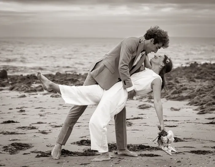 Dorine en combinaison de mariée blanche, cambrée dans les bras de son marié sur une plage rocheuse, en noir et blanc