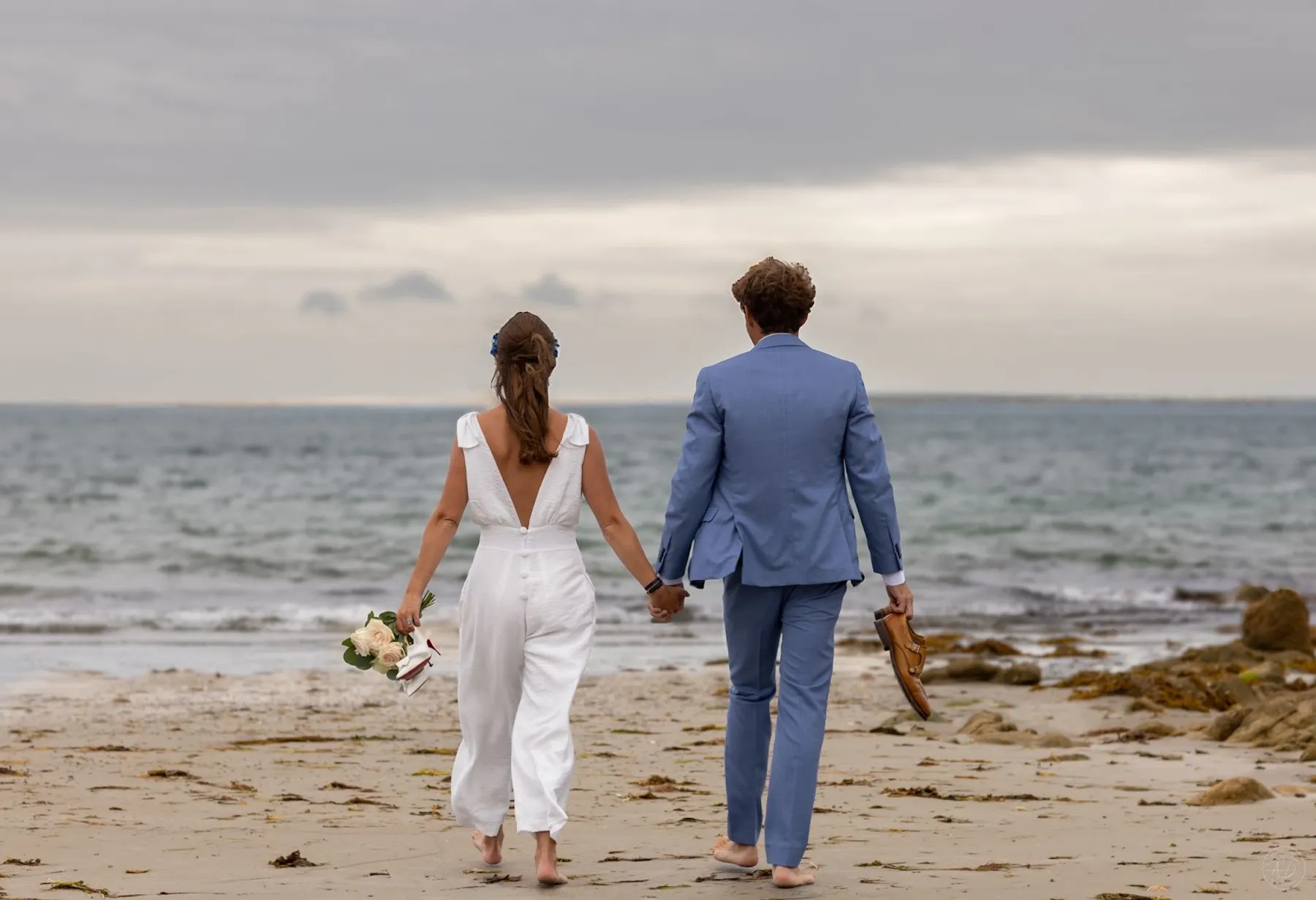 Mariés main dans la main sur la plage, de dos ; Dorine porte une combinaison-pantalon blanche au dos en V et boutons couverts