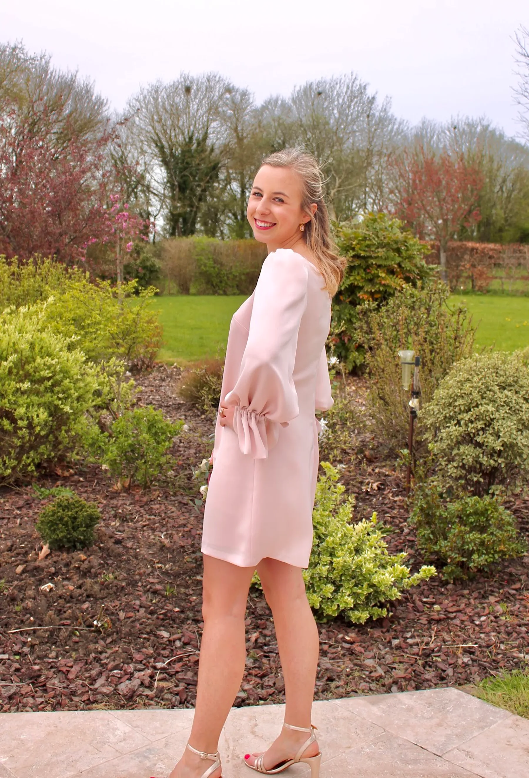 Edmée de dos dans un jardin, robe d'invitée courte rose poudré à manches bouffantes et poignets volantés