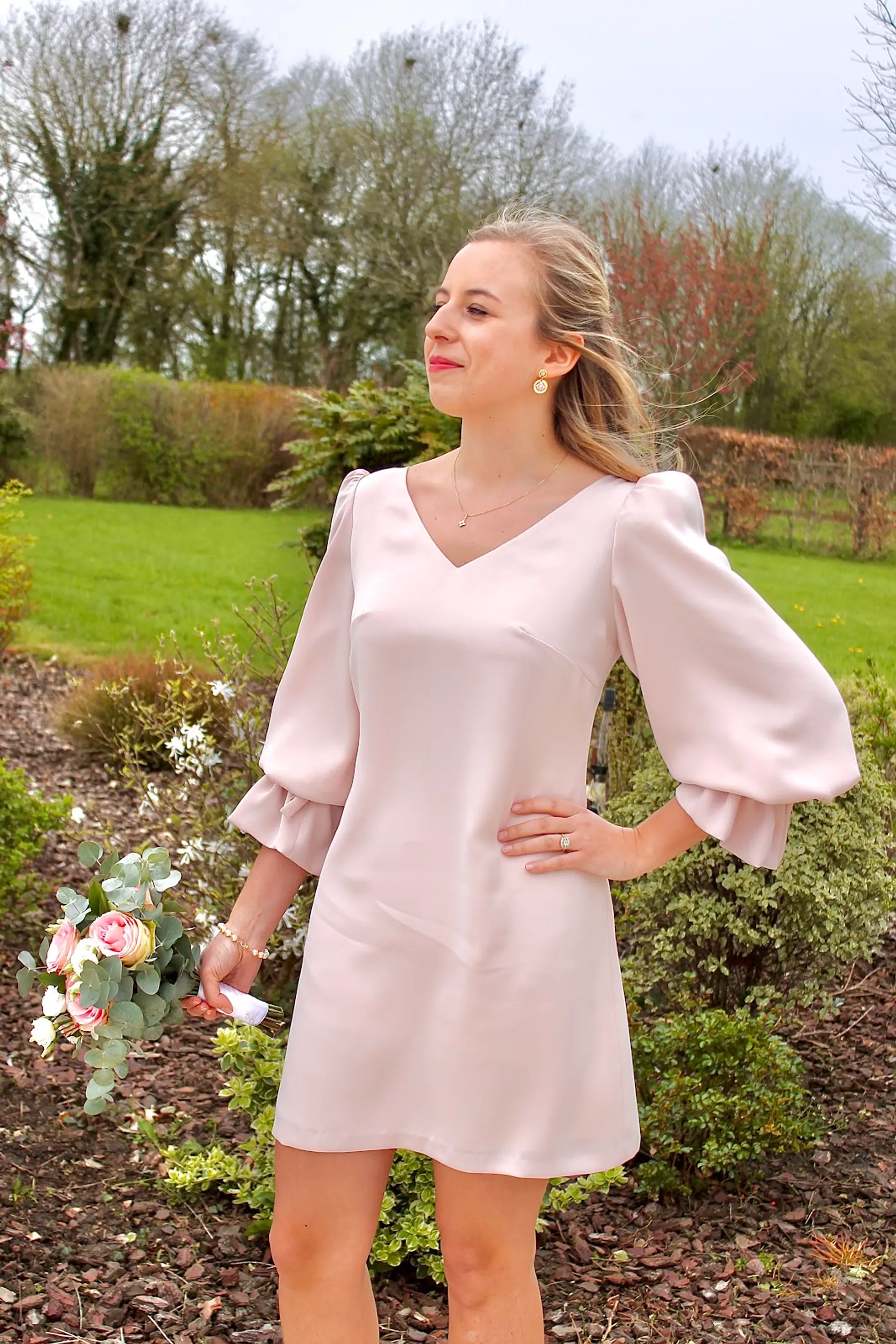 Edmée, la main sur la hanche, en robe trapèze rose poudré à col V et manches bouffantes, bouquet de roses à la main