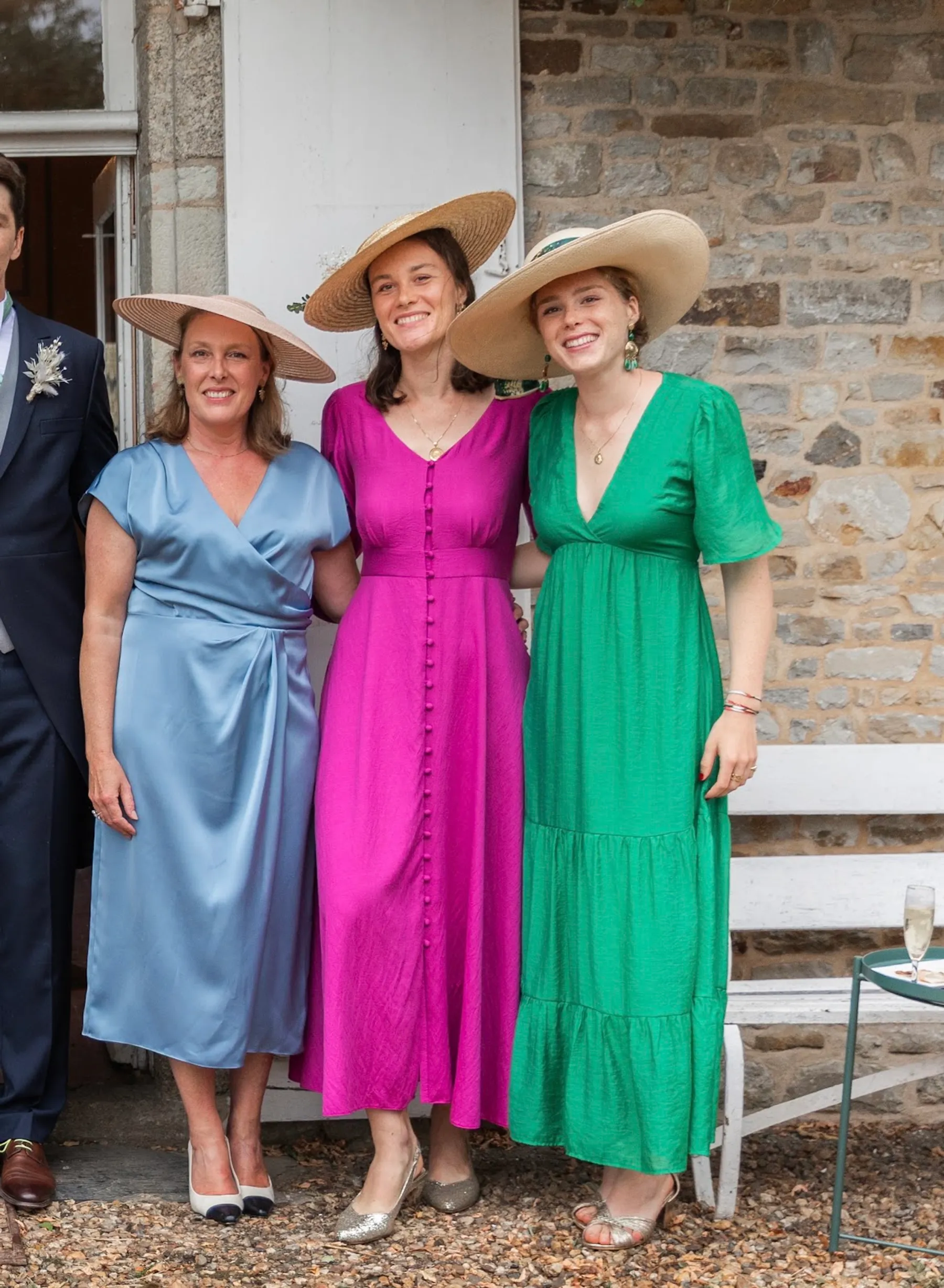 Trois invitées en chapeaux de paille devant une maison en pierre ; Éléonore, au centre, porte une longue robe fuchsia boutonnée