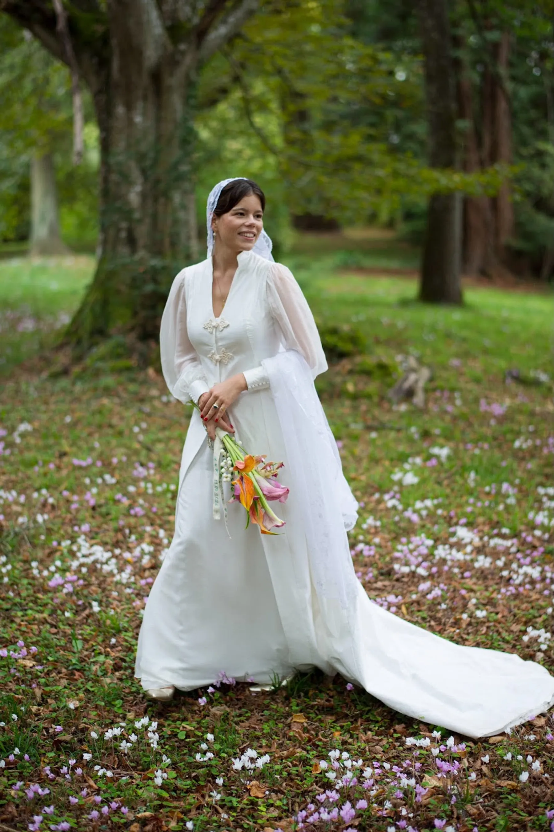 Eugénie en robe de mariée sur mesure à longues manches transparentes, traîne et voile en dentelle, bouquet de lys orangés, sous les arbres d'un parc