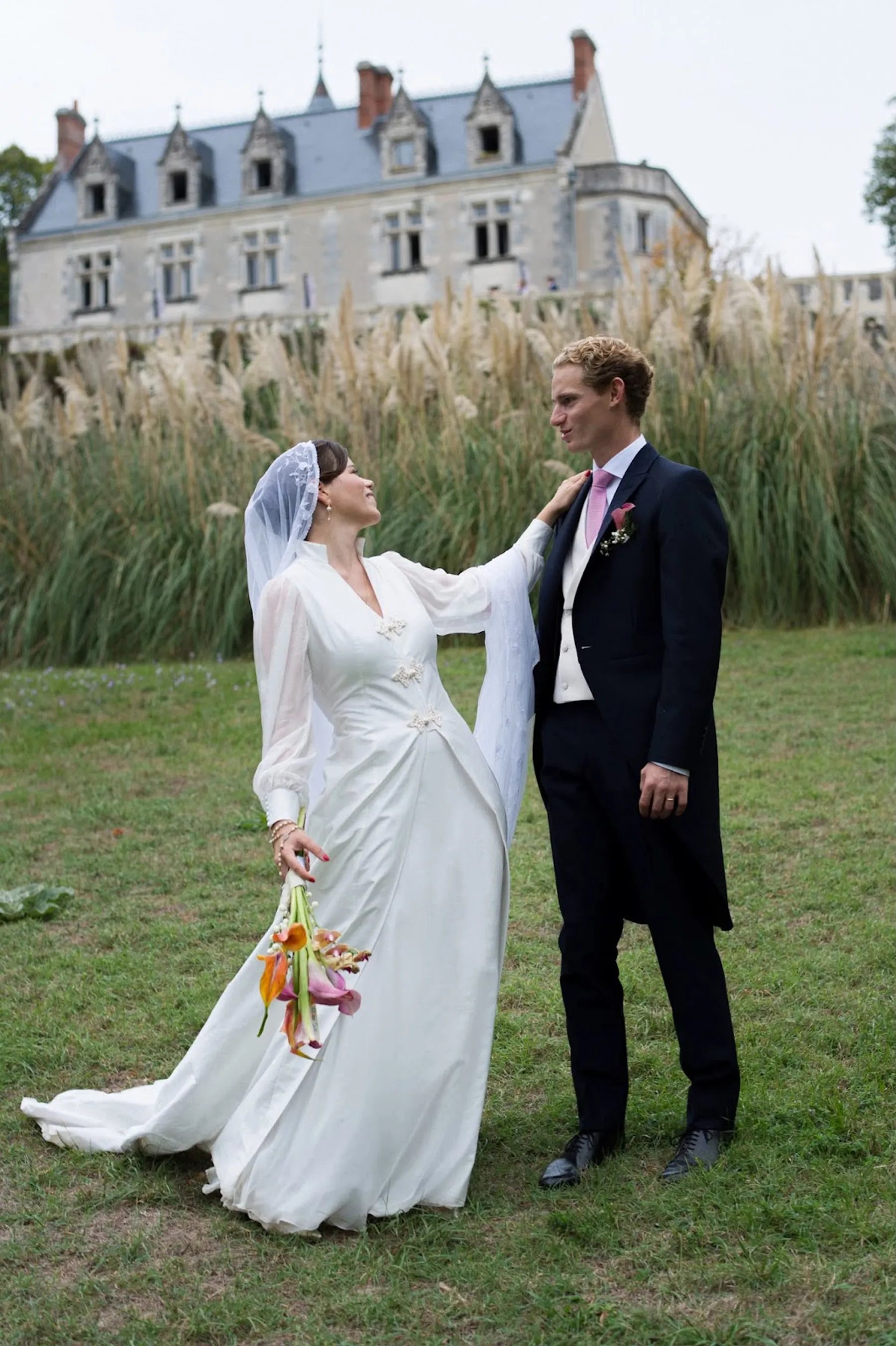 Eugénie et son marié devant un château, robe de mariée à manches en voile, longue jupe fluide et voile de dentelle