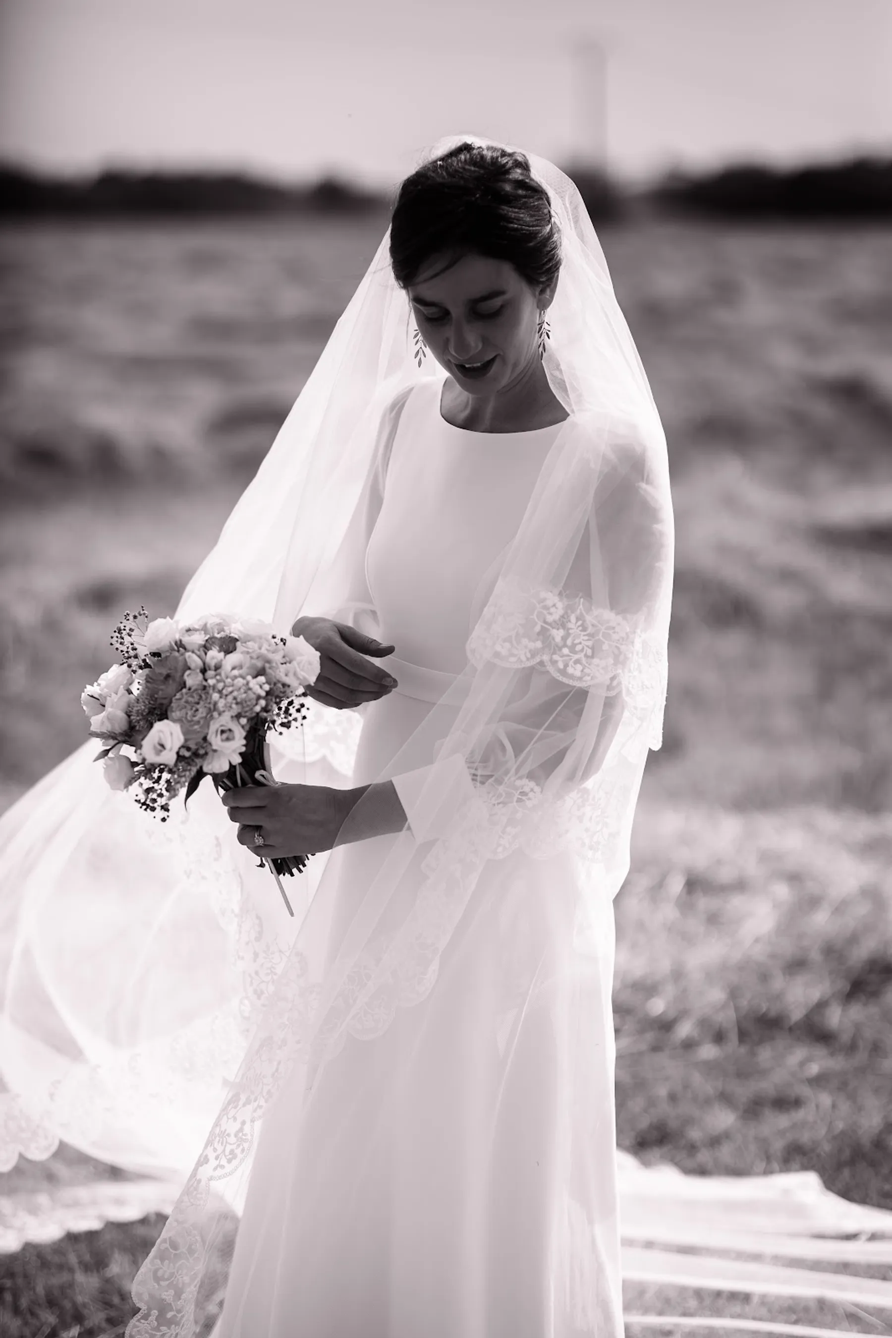Mariée en noir et blanc dans un champ, robe à col rond et manches longues sous un long voile bordé de dentelle, bouquet en main
