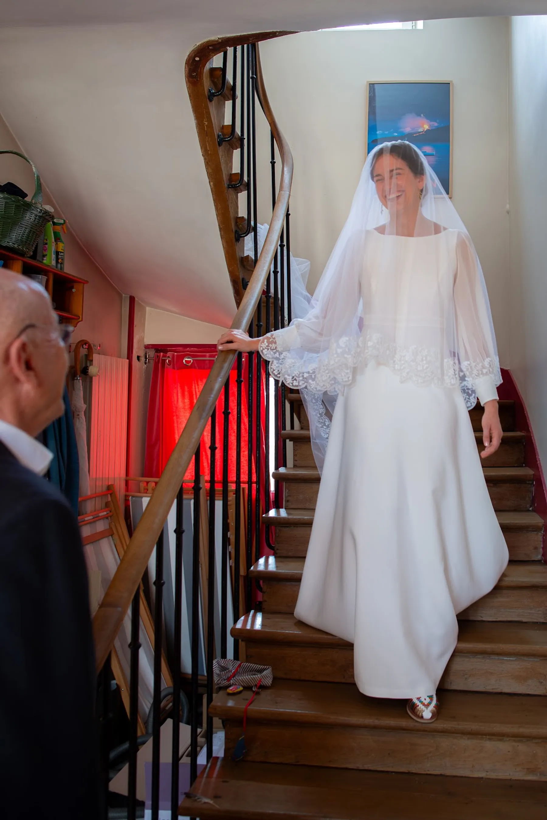 Hosanne descend un escalier en bois, voile et manches bordés de dentelle, sous le regard d'un proche