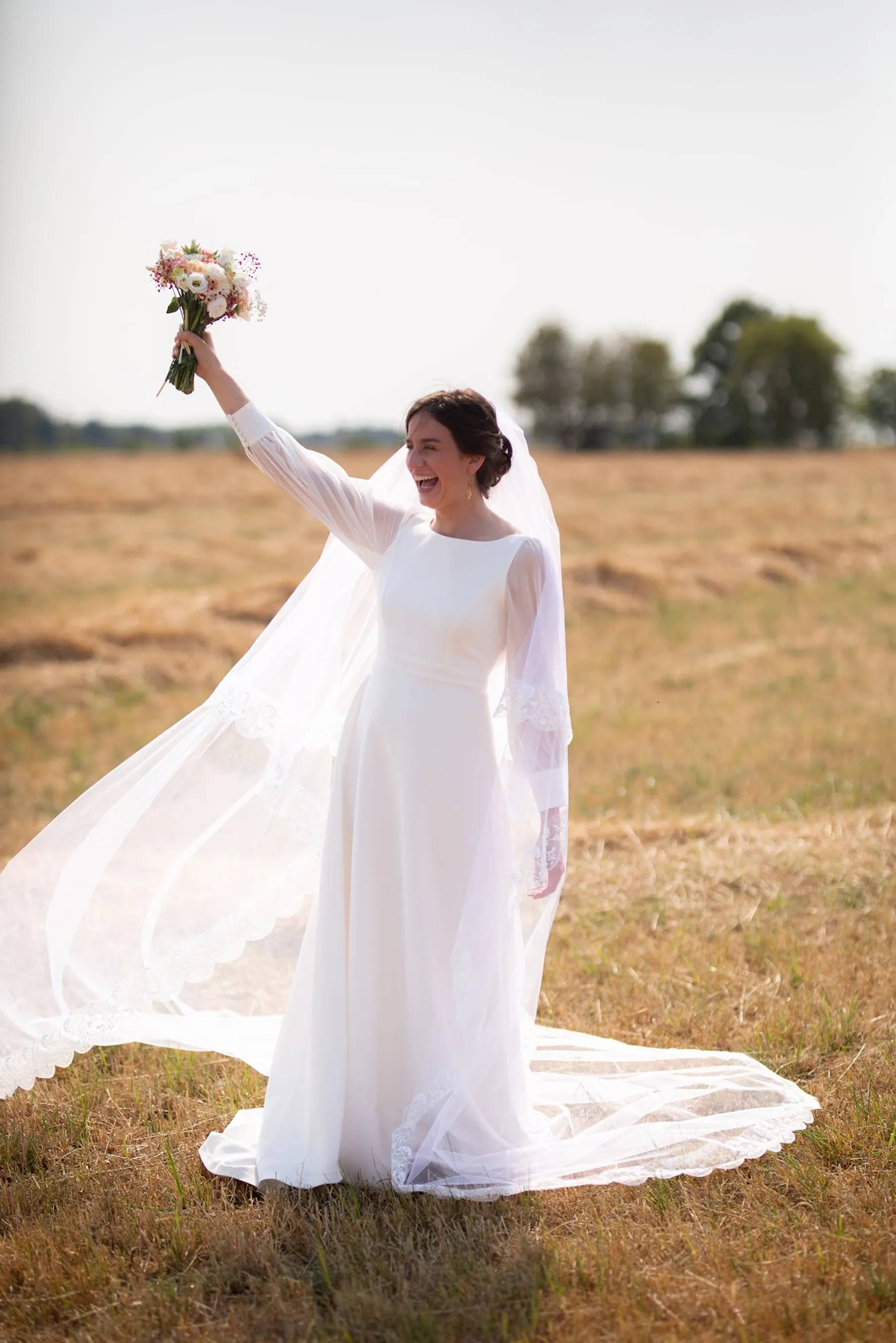 Hosanne dans un champ, un bras levé avec son bouquet, robe de mariée à col rond et manches longues, long voile flottant au vent