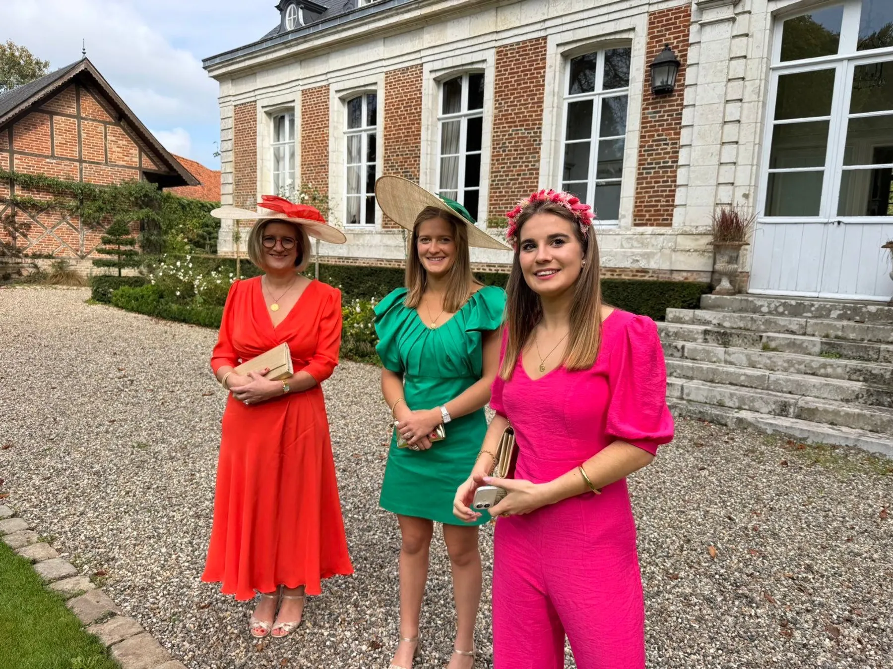 Jeanne, couronne de fleurs dans les cheveux, en combinaison fuchsia à manches bouffantes, avec deux invitées devant un château