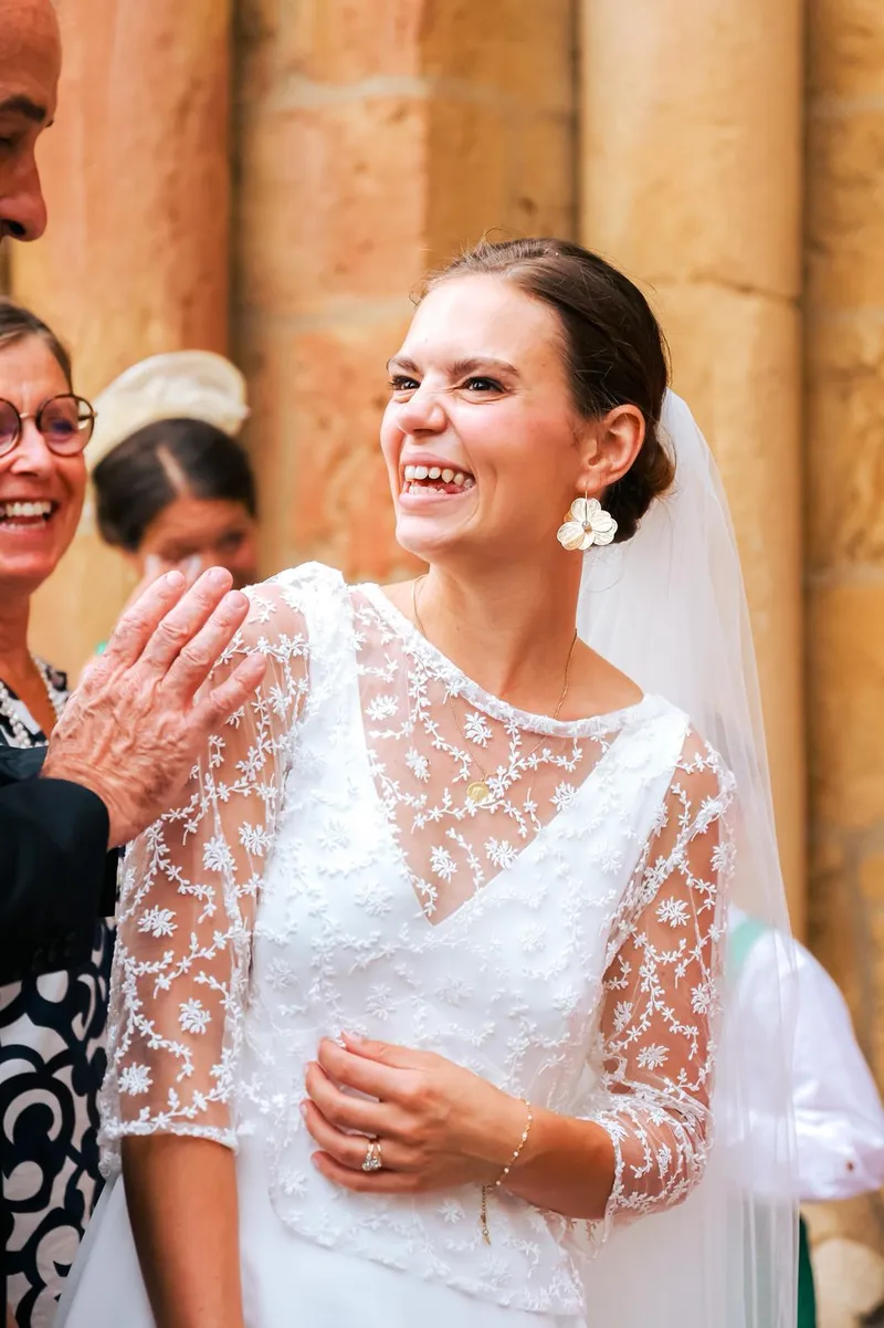 Louise éclate de rire, robe de mariée au haut en dentelle brodée de fleurs, manches longues transparentes, boucles d'oreilles fleurs