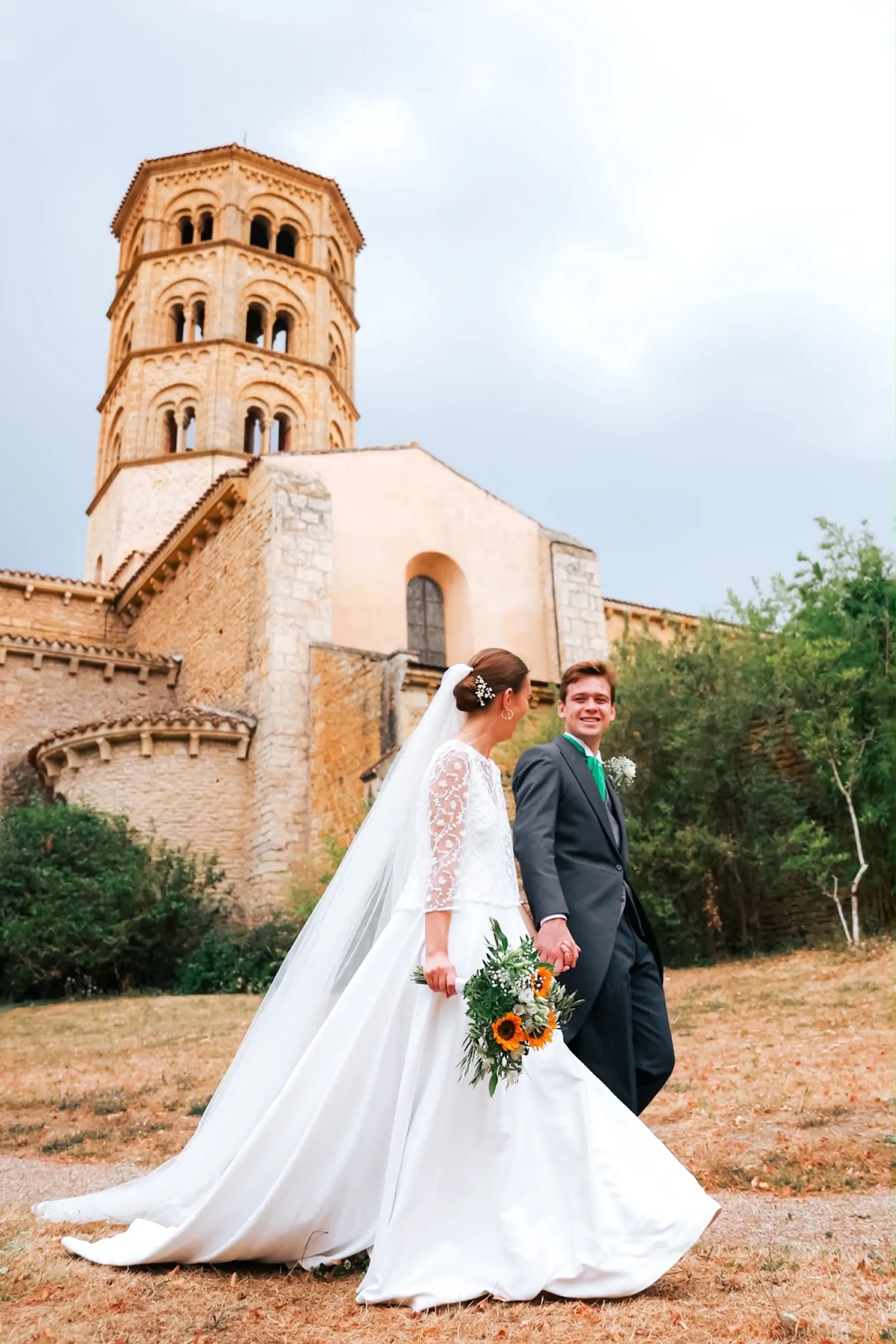 Louise et son marié main dans la main devant un clocher roman, robe à haut brodé de dentelle florale et long voile