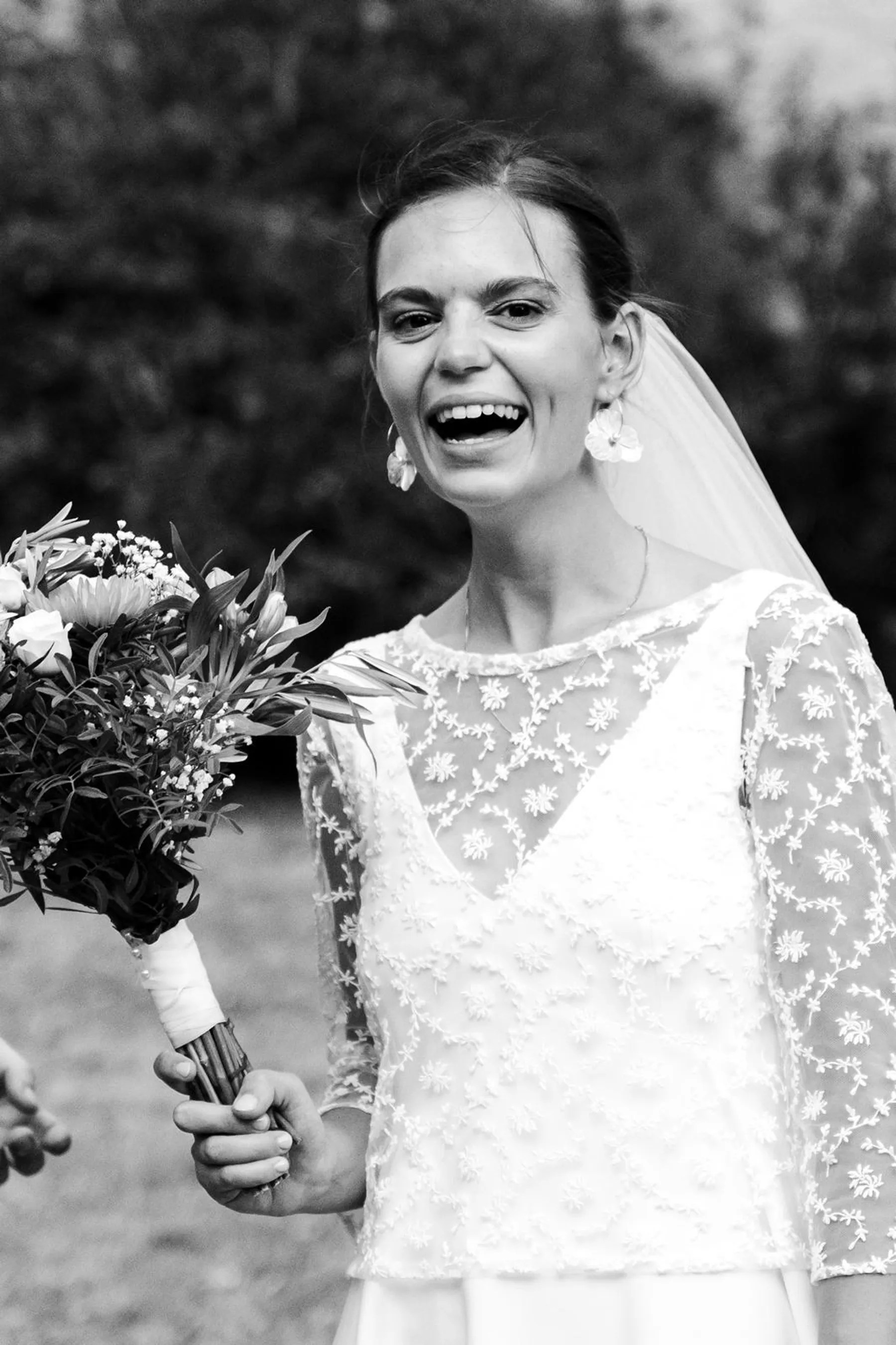 Gros plan en noir et blanc sur le haut de la robe de Louise : dentelle brodée de motifs floraux, manches longues, bouquet en main