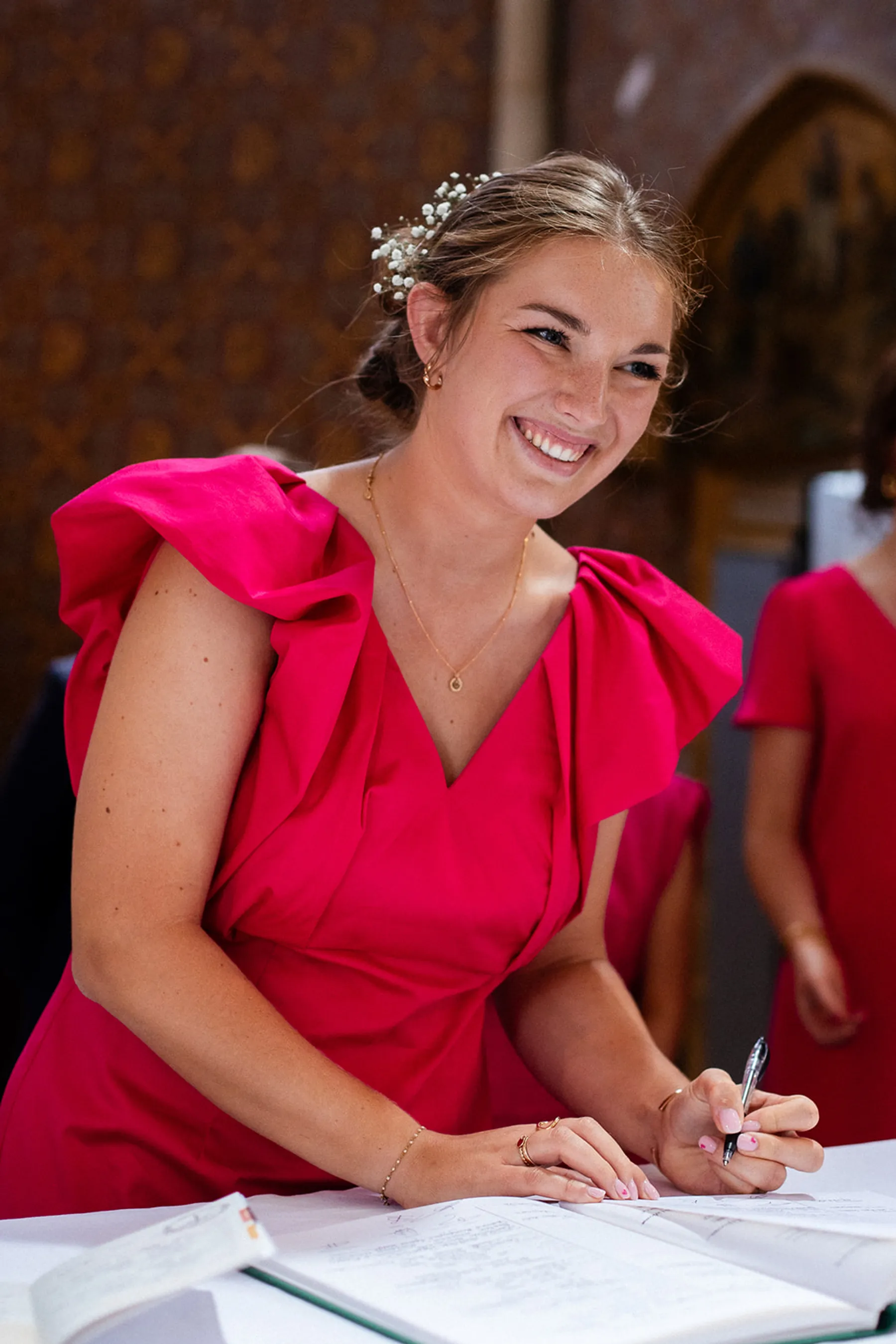 Mathilde signe le registre en souriant, robe d'invitée fuchsia à épaules à volants, petites fleurs blanches dans le chignon