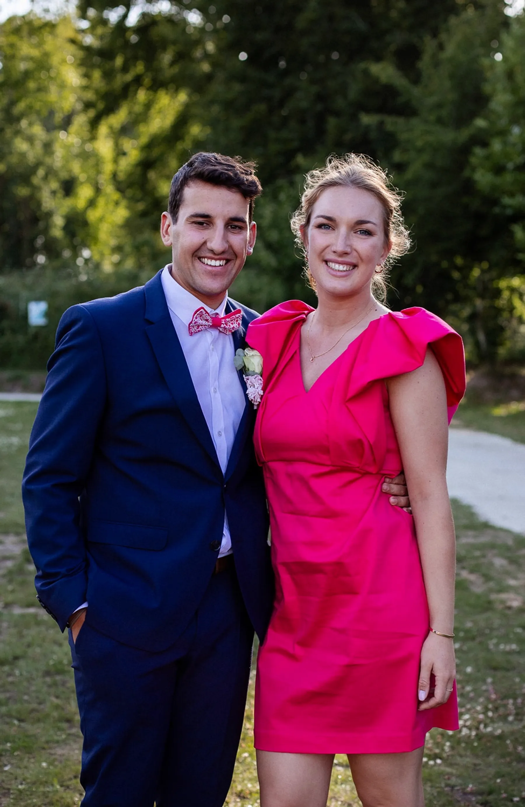 Mathilde pose avec un invité en costume bleu marine, robe fuchsia courte à volants aux épaules