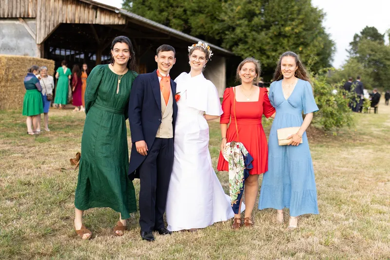 Sarah, en robe rouge à col carré et manches bouffantes, pose avec des invités à côté des mariés dans un champ