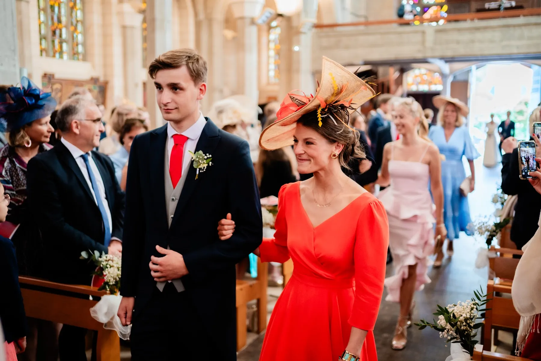Tiphaine, en robe orangée à col V et manches longues et chapeau à plumes, entre dans l'église au bras d'un invité